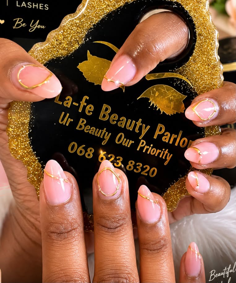 Short almond nails in sheer pink with fine gold wire outlines, held over the La-fe Beauty Parlor sign